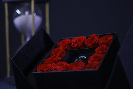 A black keepsake box with red preserved roses arranged inside, placed on a surface with a blurred background.